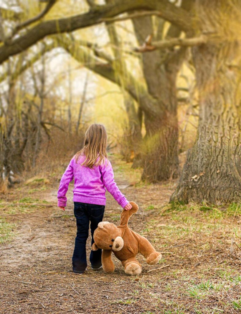 Little girl carrying her teddy bear walking down a path.